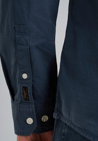 Navyblaue Baumwollhemd mit einer geknöpften Manschette und hellen Knöpfen sowie einem gewebten Etikett, das eine glatte Textur und ein maßgeschneidertes Design zeigt.