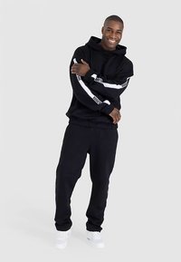 Schwarzer Kapuzenpullover mit weißen Streifen und Logo auf den Ärmeln, kombiniert mit passenden schwarzen Jogginghosen und weißen Sneakern. Glatter Stoff, lockere Passform.