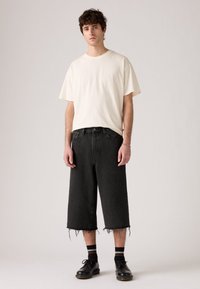 Svarte denim culotter med frynsete hem, kombinert med en kremfarget t-skjorte med korte ermer. Modellen har på seg svarte sko og stripete sokker. Enkel bakgrunn.