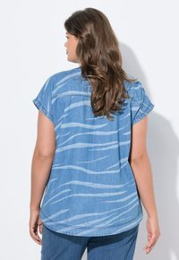 Camicia in denim azzurro chiaro con maniche corte, orlo arrotondato e strisce ondulate bianche. Texture morbida e vestibilità casual, abbinata a jeans blu.