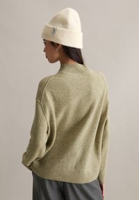 Pull tricot vert avec un col montant, des épaules tombantes et des poignets côtelés. Associé à un bonnet crème avec une étiquette. Texture douce.
