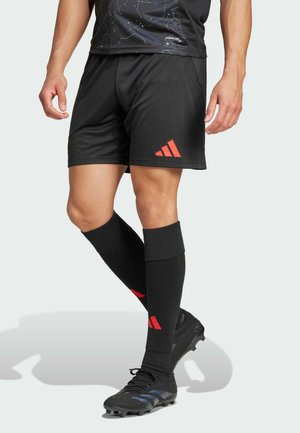 Schwarze Sportshorts mit einem roten Adidas-Logo, kombiniert mit knielangen schwarzen Socken mit roten Akzenten. Das Outfit umfasst schwarze Fußballschuhe.