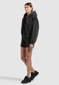 Frau steht und trägt eine schwarze Kapuzen-Denimjacke, ein schwarzes Oberteil, schwarze Denim-Shorts und schwarze Sneakers vor einfarbigem Hintergrund.