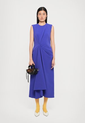 Vestito senza maniche blu cobalto con drappeggi intricati, abbinato a collant gialli e scarpe bianche a punta. Piccola borsa nera con stampa leopardata.