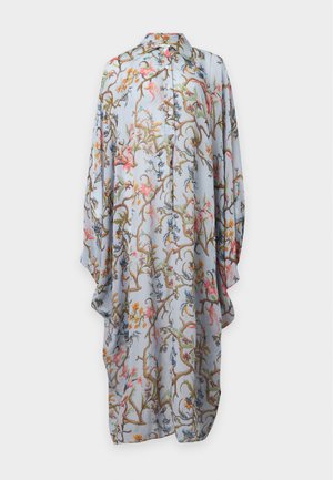 Donna con capelli lunghi e scuri che indossa un lungo vestito azzurro chiaro a fiori, ampio, e tacchi aperti neri, in piedi su sfondo bianco.