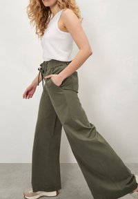 Pantalones de pierna ancha en algodón verde oliva con cintura elástica y cordón, que cuentan con bolsillos laterales y un ajuste holgado. Llevado con una camiseta sin mangas de canalé blanca.