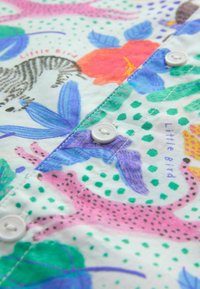 Camicia a maniche lunghe colorata con un motivo giocoso di animali e fiori su uno sfondo chiaro. I bottoni sono bianchi; il tessuto appare morbido.