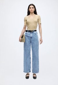 Γυναίκα που φοράει μπεζ κοντομάνικη μπλούζα, ανοιχτά μπλε τζιν με ψηλή μέση και φαρδιά πόδια, μαύρη ζώνη, μαύρα παπούτσια, κρατώντας μια μπεζ clutch με υφή.