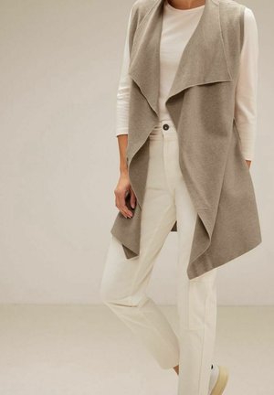 Vrouw draagt een beige mouwloos open cardigan over een wit shirt met lange mouwen en off-white broek, staande tegen een effen achtergrond.