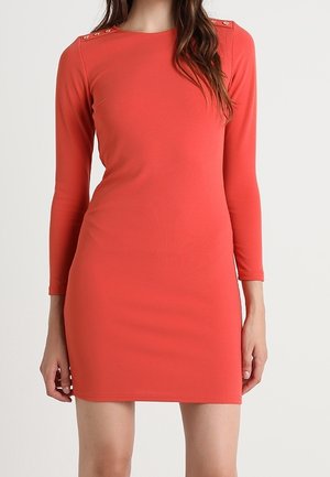 Kobieta w dopasowanej koralowej sukience z długim rękawem, z guzikami na ramionach, stojąca na tle jasnego, jednolitego tła.