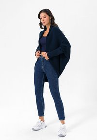 Tumesinine kudumine kardigan, laia lõikega, pikkade varrukatega ja ribadena, mille all on tumesinine lühikeste varrukatega topp ning seljas sinised kitsad teksad koos spordijalatsitega.