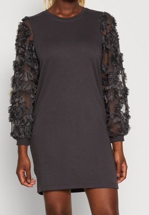 Robe courte noire avec manches longues en tulle transparent ornées d'appliqués floraux texturés, col rond, portée par une personne à la peau marron moyen.