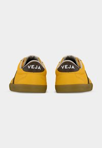 Κίτρινα αθλητικά παπούτσια με καφέ λεπτομέρειες στη φτέρνα, με στρογγυλεμένο σχήμα, υφασμένα λαστιχένια πέλματα και λευκή επιγραφή "VEJA" στο πίσω μέρος.