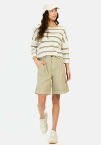 Leichtes, gestreiftes Langarmoberteil in Creme und Taupe, kombiniert mit beigen Shorts mit umgeschlagenen Säumen. Dazu werden weiße Sneakers und eine Cap getragen.