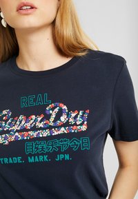 Marinblå bomullströja med färgglada blommotiv och grön text som säger "REAL SuperDry", med rund halsringning och korta ärmar.