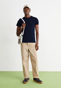 Tricou bleumarin cu buzunar pe piept, pantaloni bej și bocanci de drumeție bleumarin. Capac de un bej deschis. Stând pe o podea verde.