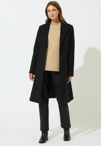 Cappotto di lana nera con una vestibilità sartoriale, maniche lunghe, revers a punta e due bottoni, abbinato a un maglione beige e pantaloni neri.
