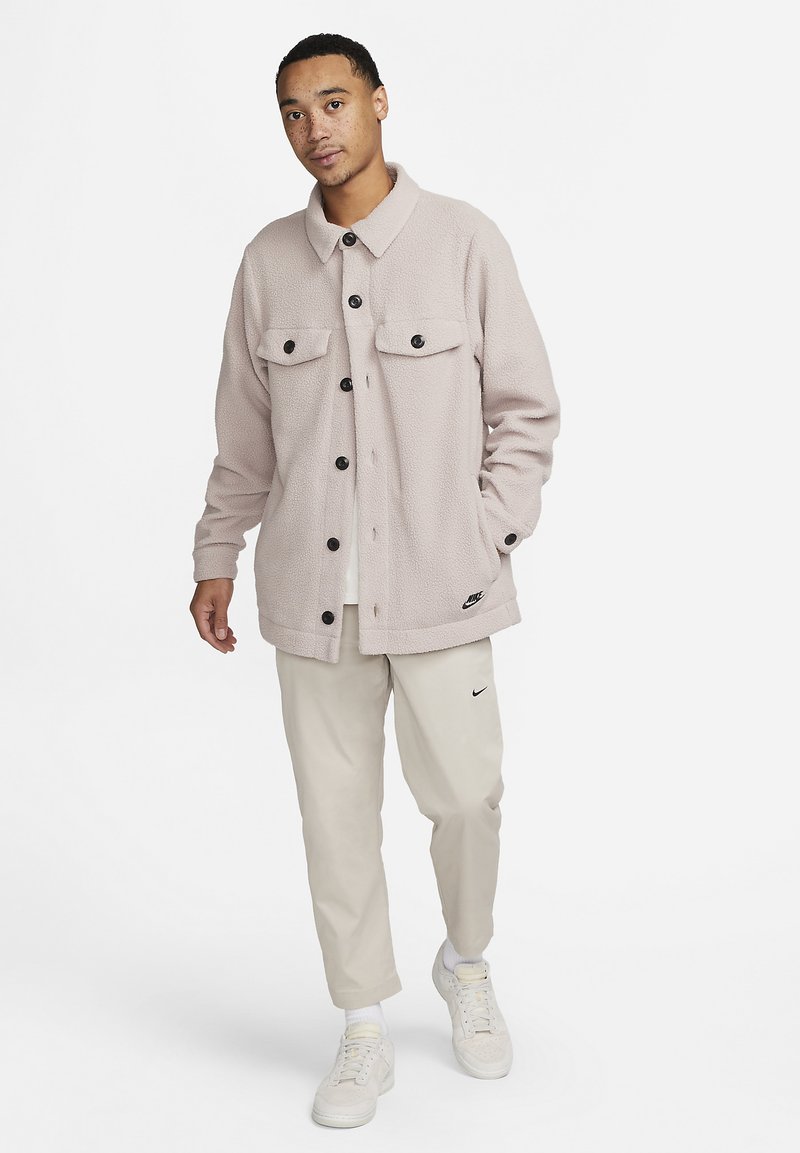 Lys rosa fleecejakke med to brystlommer, sorte knapper og et Nike-logo. Kombinert med lyse beige bukser og hvite joggesko.