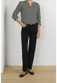 Sort og hvid mønstret bluse med v-udskæring og trekvartærmer, parret med højtaljede sorte bukser og sorte flade sko.