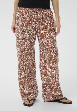 Femme portant un pantalon à jambes larges à motif floral orange et marron, des tongs noires et des bijoux en or, debout devant un fond uni.