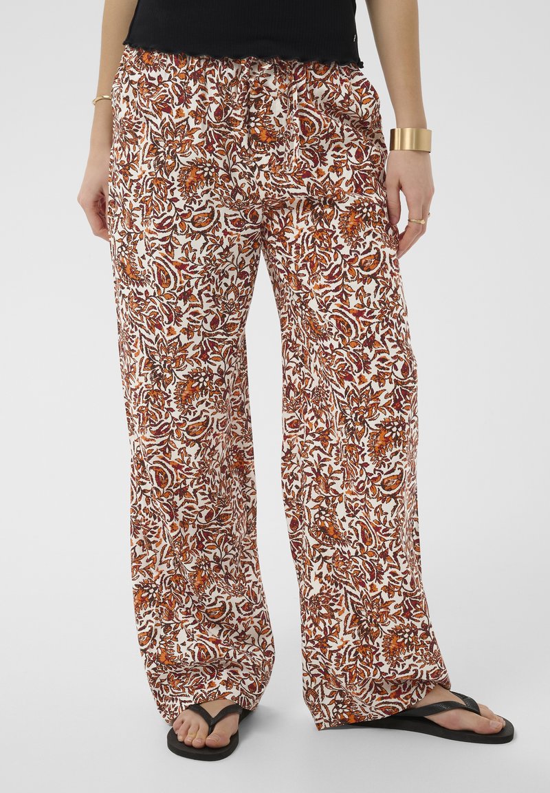 Frau trägt weit geschnittene Hosen mit orange-braunem Blumenmuster, schwarze Flip-Flops und Goldschmuck, steht vor einfachem Hintergrund.