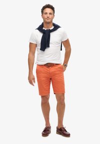 Biała koszulka, pomarańczowe szorty, granatowy sweter związany na szyi, brązowy pasek, brązowe buty. Model stoi z neutralnym wyrazem twarzy na białym tle.