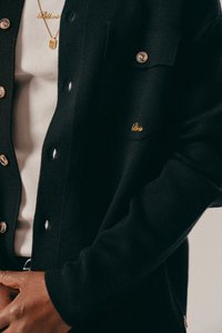 Close-up van een persoon die een zwarte knoopcardigan draagt boven een beige geribd shirt, met een gouden ketting en geborduurde tekst op de zak van de cardigan.