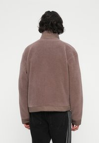 Oseba s srednje dolgimi lasmi, oblečena v mauve sherpa flis jakno in črne hlače z stranskimi črtami, stoji proti beli ozadju, pogled od zadaj.