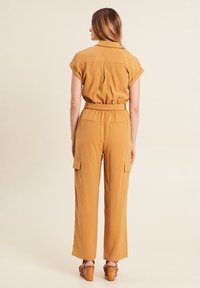 Mosterdgele jumpsuit met korte opgerolde mouwen, elastische tailleband en zijcargo zakken. Gladde textuur en recht model.