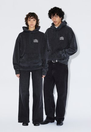 Twee jonge volwassenen staan zij aan zij, gekleed in zwarte oversized hoodies met de tekst "Utopia" en zwarte broeken tegen een effen witte achtergrond.