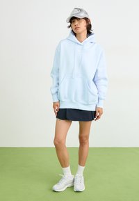 Hoodie azul claro com um bolso frontal e cordão, combinado com uma saia mini preta e ténis brancos. Boné cinza com detalhe do logotipo.