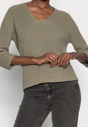 Person iført en lys olivengrøn ribstrikket sweater med trekvartlange ærmer og mørkegrå højtaljede jeans, hænderne foldet.