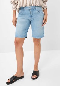 Shorts en denim bleu clair avec une coupe décontractée, design à cinq poches et fermeture à bouton visible, assortis à des sandales noires à brides croisées.