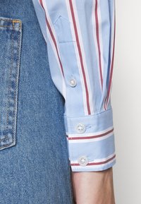 Manica di camicia a righe azzurre chiare con righe verticali rosse e bianche, chiusura a bottoni e polsino a punta, abbinata a un tessuto in denim.
