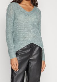 Ljus teal stickad tröja med V-hals, långa ärmar och en lätt böjd fåll, i kombination med svarta skinnbyxor. Mjuk textur och löst sittande.