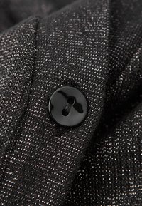 Černý lesklý dvouotvorový knoflík přišitý na texturované černobílé tkanině, pohled z blízkosti.