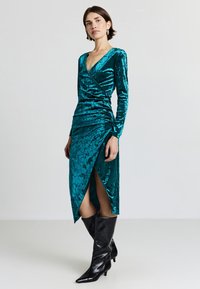 Robe en velours turquoise à manches longues et décolleté en V, avec un design froncé et une fente haute mi-cuisse, accompagnée de bottes noires montant jusqu'aux genoux.