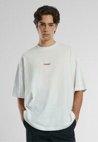 T-shirt blanc surdimensionné en coton, avec un col rond et des manches courtes. "Interdit" est brodé en rouge sur la poitrine.