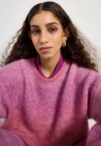 Pletený růžový svetr s přechodem odstínů, kulatým výstřihem a jemnou texturou, doplněný fialovým topem pod ním. Zlaté kruhové náušnice.