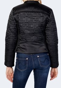 Schwarze, gesteppte Jacke mit Textur und Logomuster; glatte Schultern, taillierter Schnitt; getragen über dunklen Jeans mit auffälligem Taschendesign.
