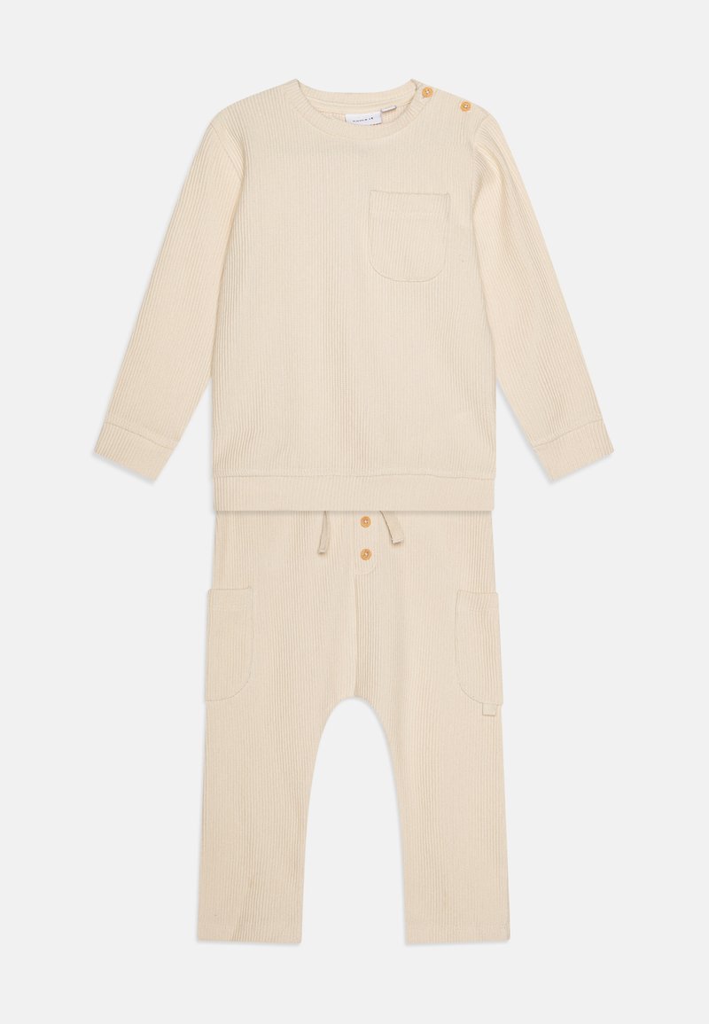 Bež kombinacija od dva dijela s dugim rukavima, džepom na prsima, prilagodljivim strukom i dva bočna džepa. Sadrži drvene kopče kao detalj.