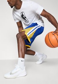 Hvit t-skjorte med svart basketball-logo, blå shorts med gule og hvite striper, hvite sneakers og en oransje basketball, som viser atletisk bevegelse.