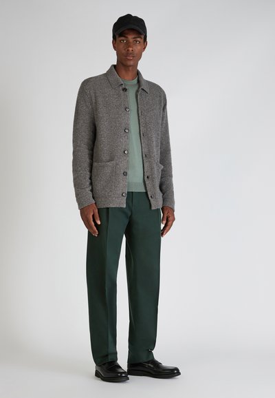 Grau wollene Strickjacke mit Fronttaschen, schwarzen Knöpfen, über einem grünen Pullover getragen. Kombiniert mit grünen Hosen und schwarzen Schuhen.