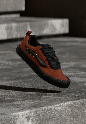 Rust-colored sneaker with black ruffled laces, black sole, and metal studs hovering above gray concrete steps.