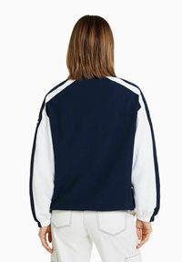 Veste bicolore bleu marine et blanc avec poignets élastiques. Présente une coupe décontractée et des surpiqûres contrastantes. Le matériau semble léger et lisse.