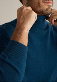 Homme barbu ajustant le col d'un pull à col roulé bleu, mettant l'accent sur les mains et la partie supérieure de la poitrine.