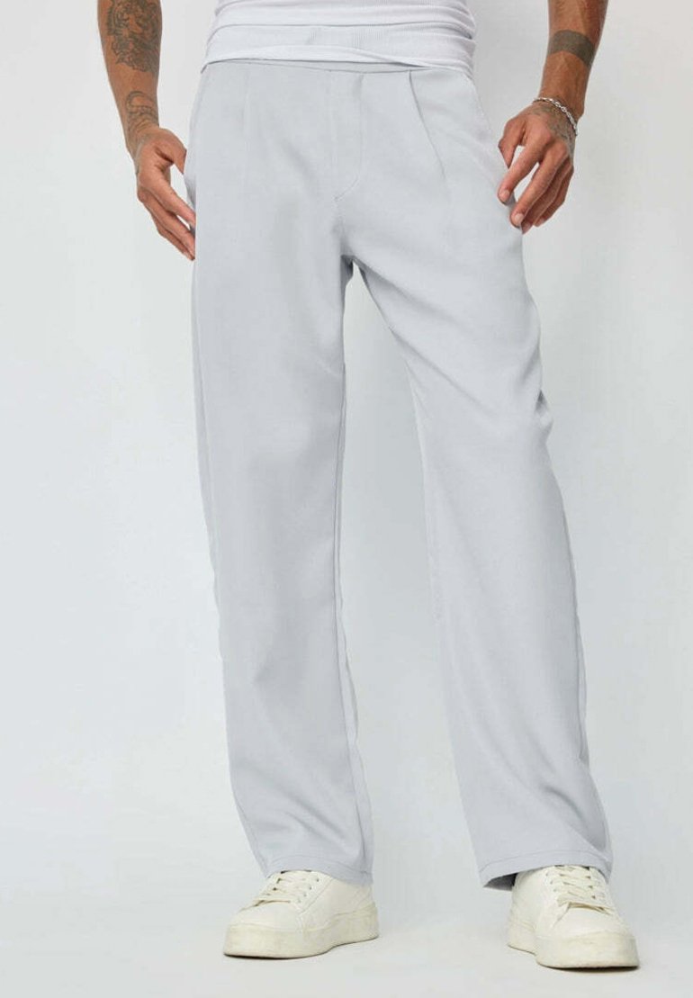 Pantalons de survêtement gris clair avec une coupe décontractée, présentant une texture lisse, des plis devant et une taille élastique, associés à des baskets blanches.