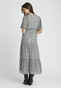 Robe maxi en tissu floral transparent avec des fleurs noires et blanches, design à étages, manches courtes et détail des boutons dans le dos. Portée avec des bottes noires.