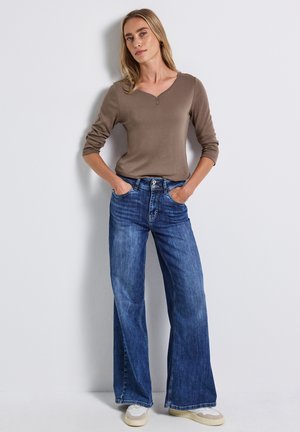 Haut marron à manches longues avec un col en V et une fermeture éclair, associé à un jean en denim bleu à jambes larges. Le jean présente un léger délavage et des coutures visibles.