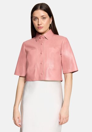 Frau trägt ein kurzes rosa Lederhemd mit Knöpfen und kurzen Ärmeln und einen weißen Rock, stehend vor schlichtem Hintergrund.